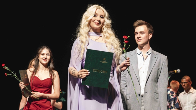 sieviete gaiši violetā kleitā tur diplomu un sarkanu ziedu, blakus stāv vīrietis pelēkā uzvalkā