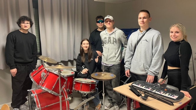 Izglītojamo grupa stāv pie bungu komplekta un sintezatora mācību telpā