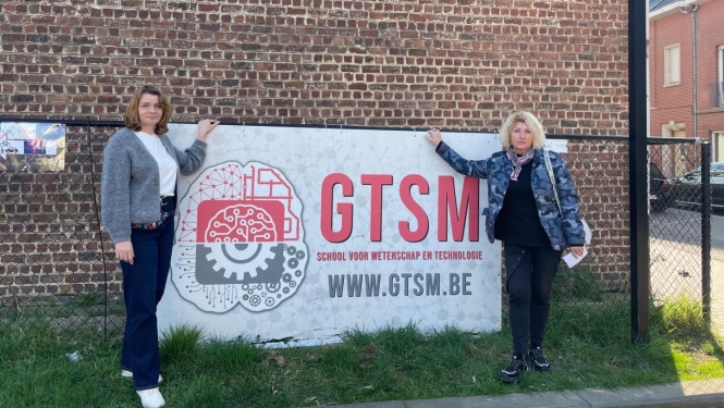 Divas pedagoģes stāv pie GTSM skolas informatīvās zīmes pie ēkas Beļģijā