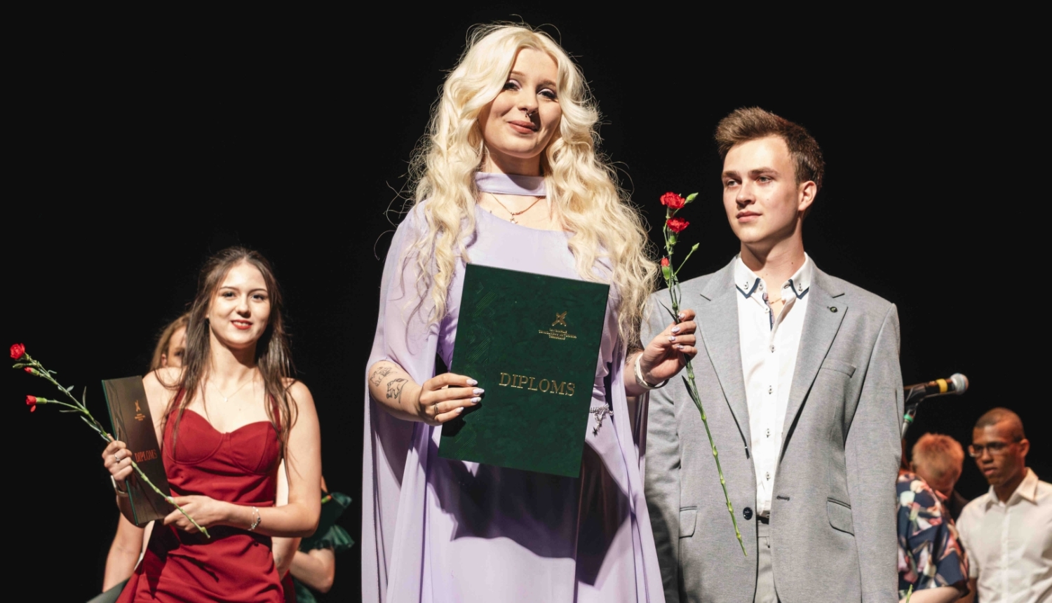 sieviete gaiši violetā kleitā tur diplomu un sarkanu ziedu, blakus stāv vīrietis pelēkā uzvalkā