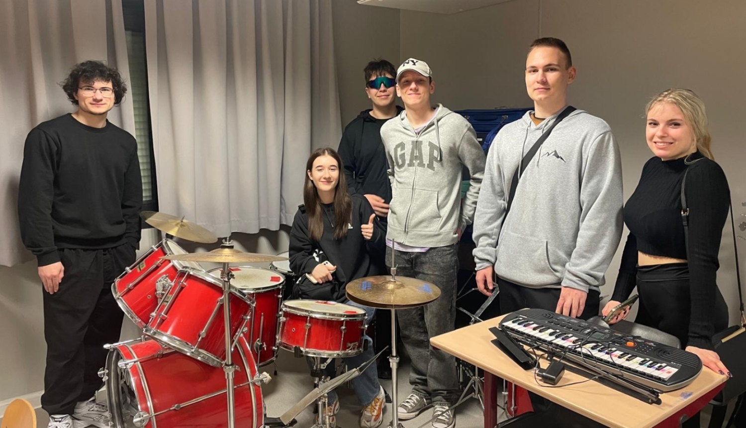 Izglītojamo grupa stāv pie bungu komplekta un sintezatora mācību telpā