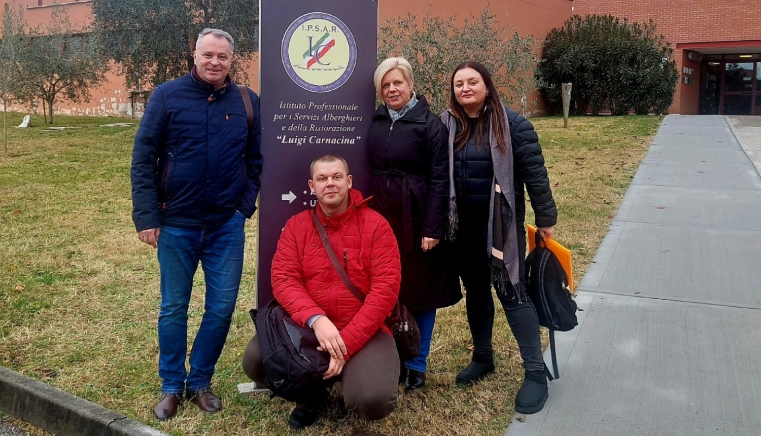 Četri tehnikuma pārstāvji pozē pie izglītības iestādes informatīvās zīmes ārpus ēkas