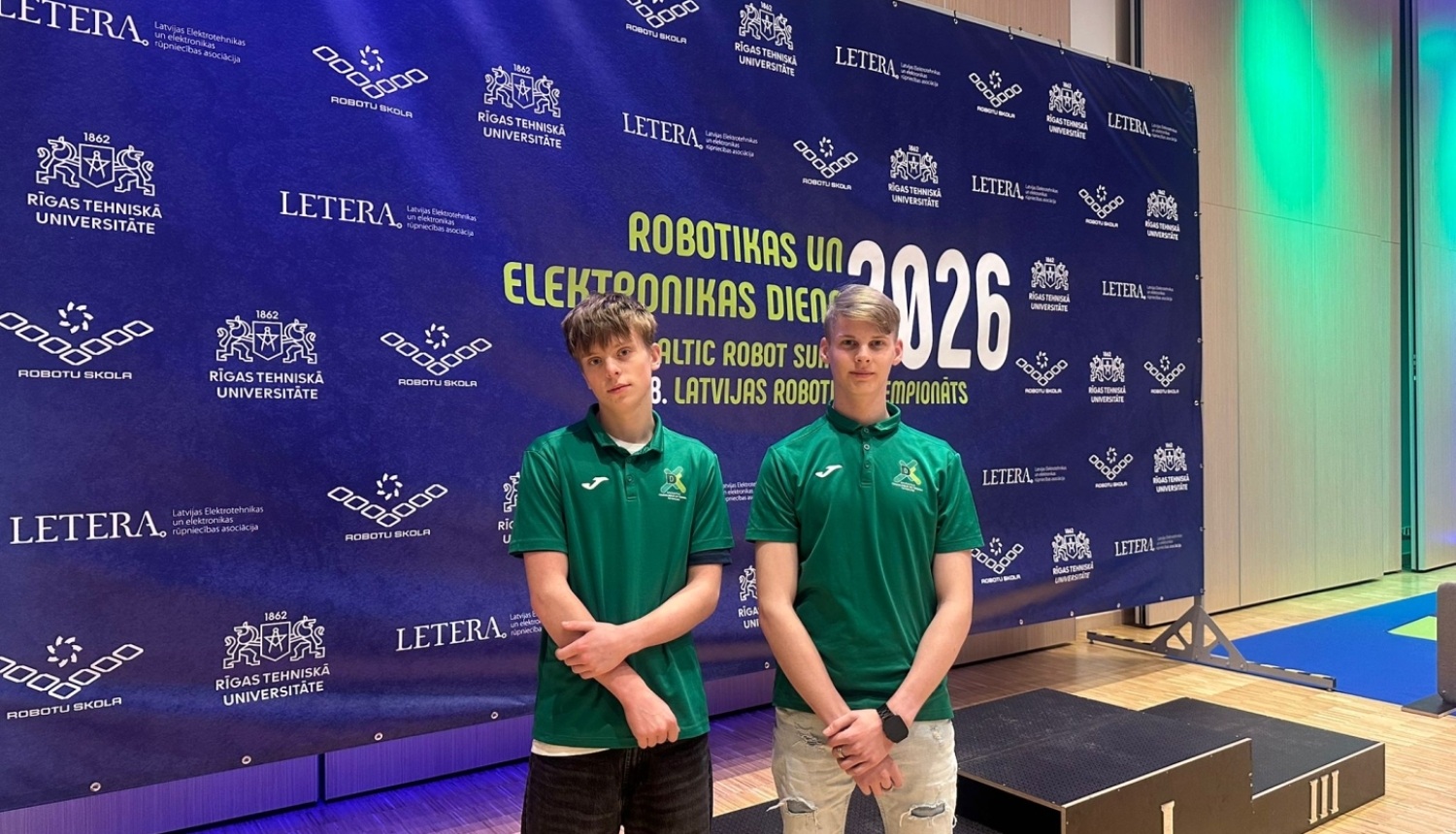 Divi jaunieši stāv pie “Robotikas un elektronikas dienas 2026” stenda