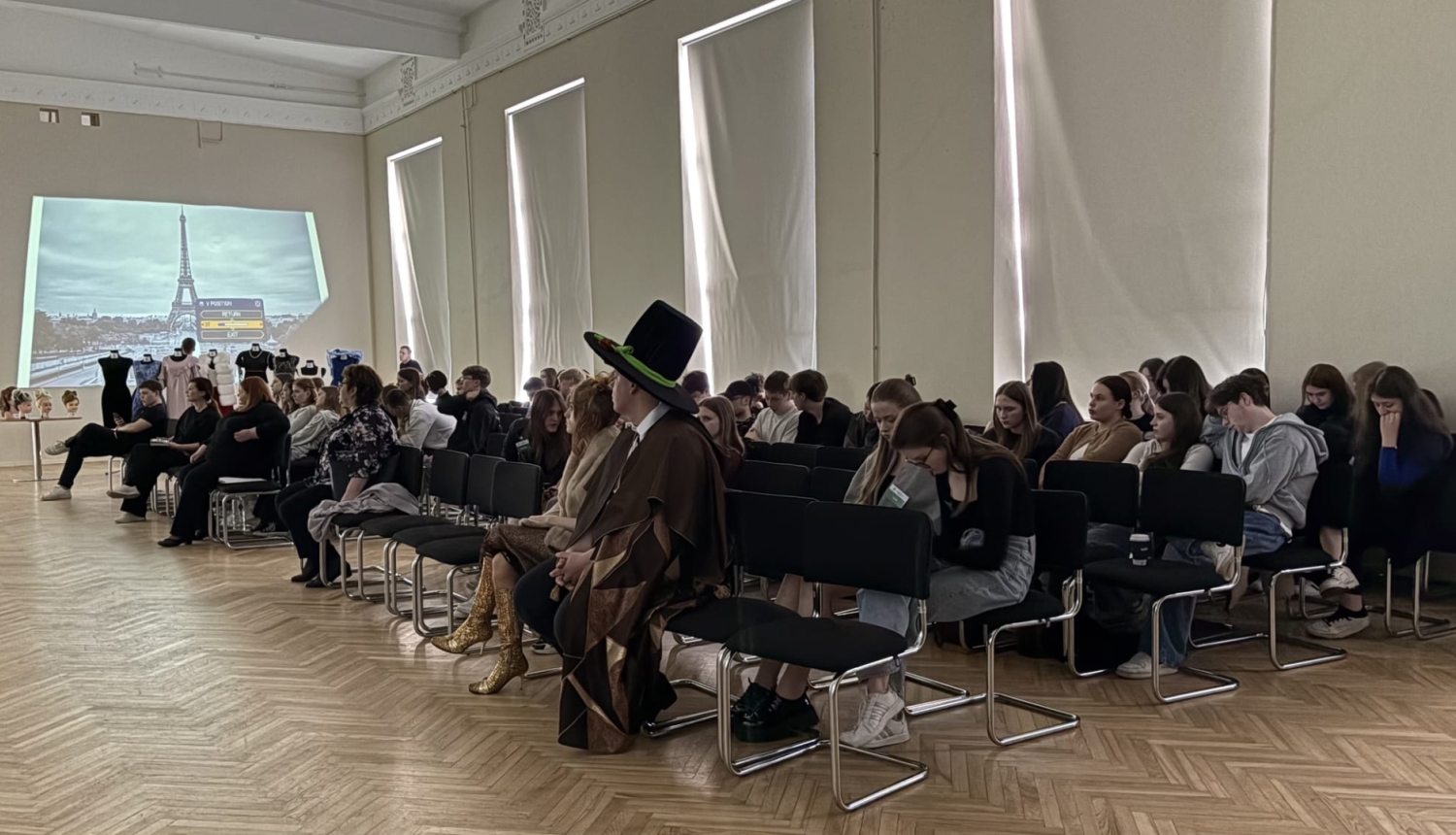 Pasākuma dalībnieki sēž zālē un vēro projekta prezentāciju
