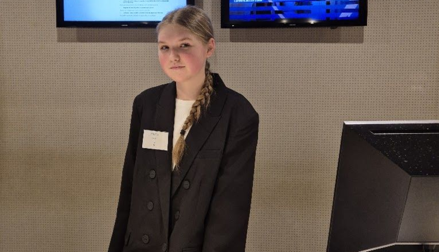     Izglītojamā stāv aiz viesnīcas reģistrācijas letes praksē Itālijā, fonā redzami divi ekrāni