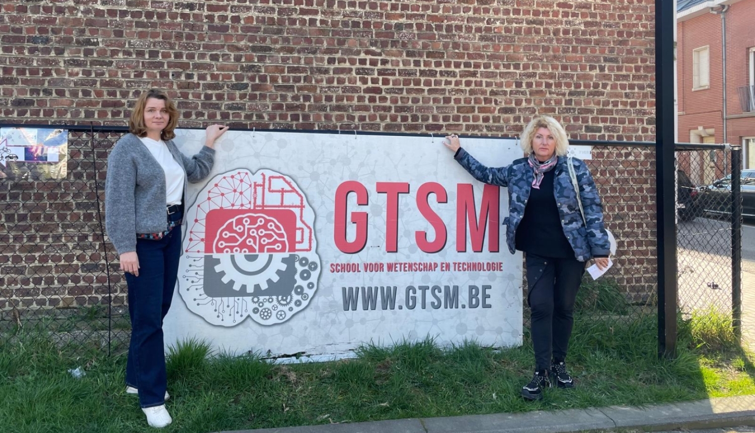 Divas pedagoģes stāv pie GTSM skolas informatīvās zīmes pie ēkas Beļģijā