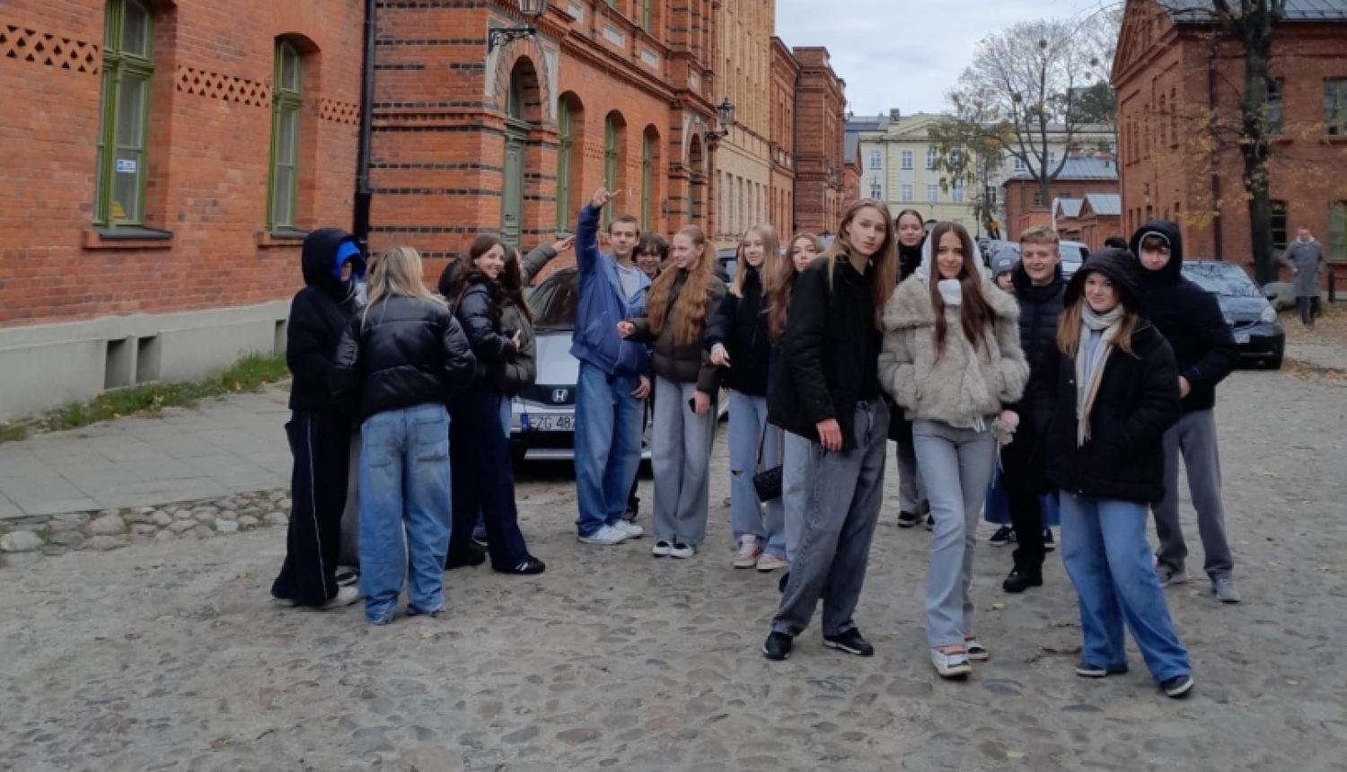 Grupa izglītojamo pozē uz bruģētas ielas Lodzā, Polijā, fonā redzamas vēsturiskas sarkano ķieģeļu ēkas.